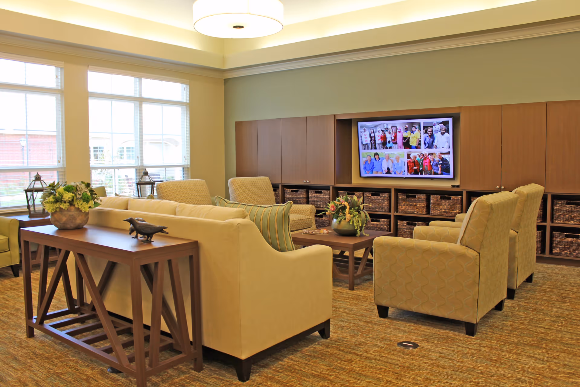A cozy living room area in a senior living facility with beige sofas and armchairs arranged around a wooden coffee table. A flat-screen TV mounted on a wall unit displays a collage of photos featuring groups of people. Large windows let in natural light, and decorative items like plants and lanterns are placed on furniture.