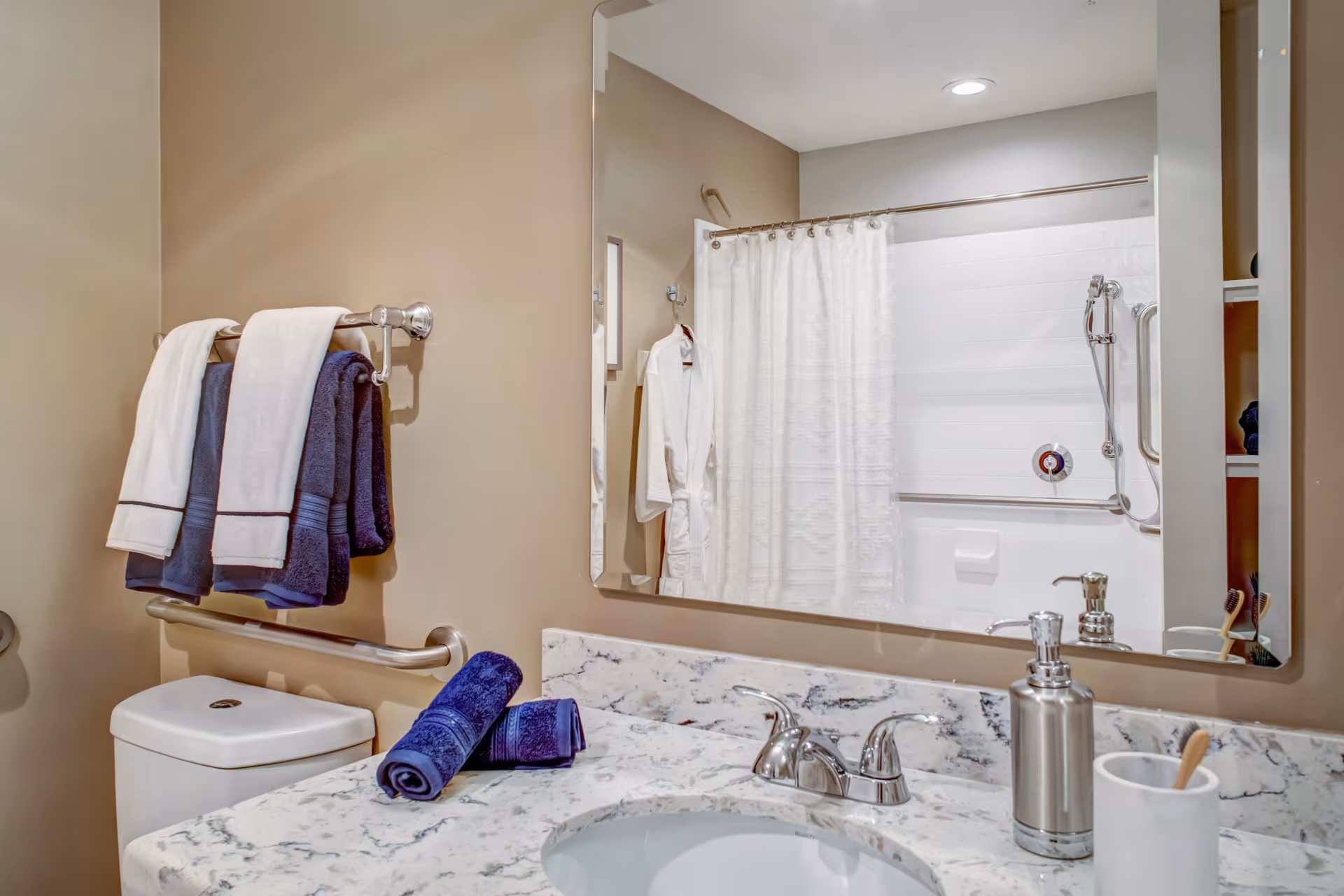 Bathroom vanity with marble countertop and sink, towels on a rack, and a mirror reflecting a shower with grab bars and a curtain.