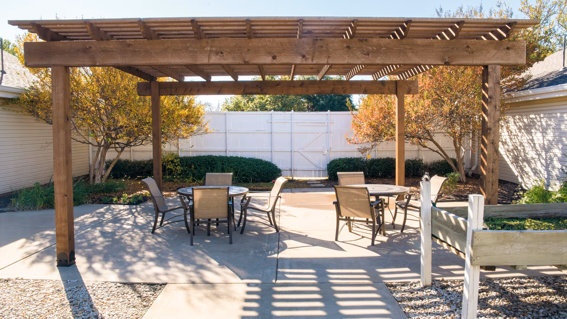 Outdoor courtyard with a wooden pergola shading round tables and chairs on a concrete patio surrounded by a white fence and landscaping.