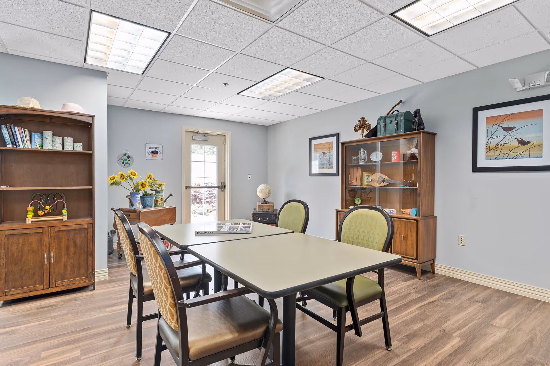 A well-lit room with a rectangular table surrounded by four chairs with patterned upholstery. The room has light gray walls and wood flooring. There are two wooden cabinets, one with shelves holding books, decorative items, and a toy, and the other with glass doors displaying various objects. Two framed pictures of birds hang on the walls. A door with a window leads outside, and a small table with a globe and a vase of sunflowers is near the door.