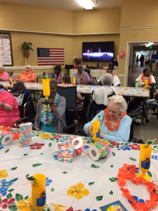 A group of elderly people seated around tables decorated with colorful floral tablecloths and tropical-themed decorations, including yellow cups with palm tree designs and leis. The room has beige walls, an American flag on the wall, and a television mounted on the wall showing a performance. Some people are wearing leis and appear to be engaged in an activity or social event.