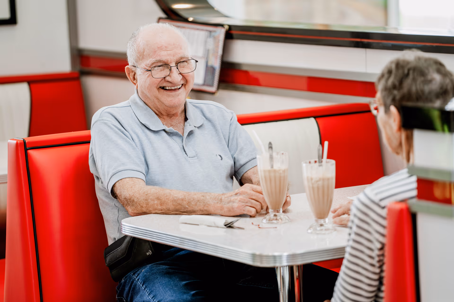 Two older adults sit in a red-and-white diner booth enjoying milkshakes, with a smiling man facing the camera.