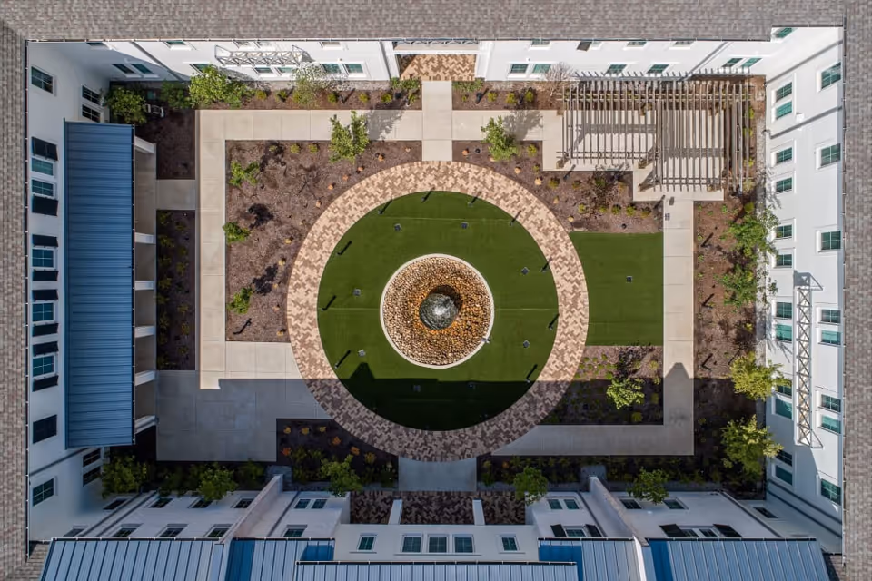 Aerial view of a courtyard at The Claiborne at Westlake featuring a circular fountain in the center surrounded by green artificial turf and a patterned brick walkway. The courtyard is enclosed by a white building with multiple windows and has landscaped areas with small trees and plants.