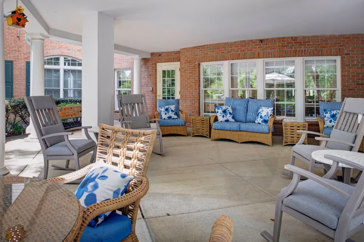 Covered outdoor patio area with wicker and cushioned seating including chairs and sofas with blue and white floral and striped pillows, surrounded by brick walls and large windows.