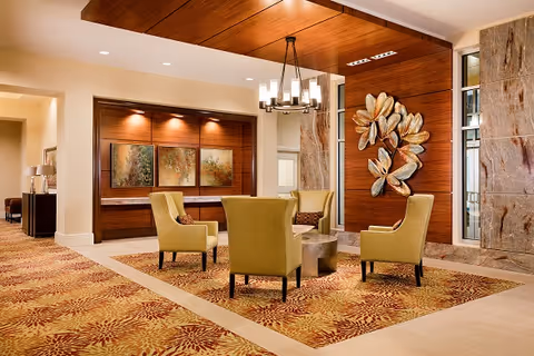 A well-lit lounge area with four green upholstered chairs arranged around a round coffee table on a patterned carpet. The walls feature wood paneling, decorative artwork including a large metallic floral sculpture, and framed paintings. A modern chandelier hangs from the ceiling, and there is a hallway visible in the background.