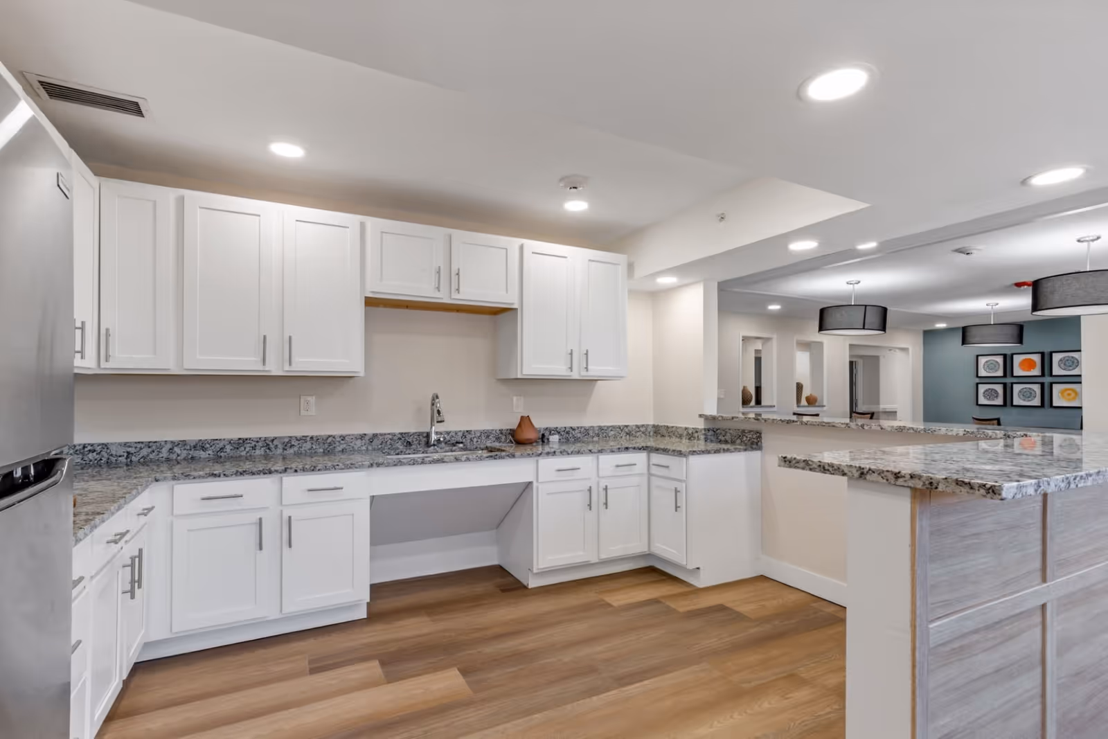 Modern kitchen area with white cabinets, granite countertops, a stainless steel refrigerator, and wood flooring. The kitchen opens to a common area with pendant lights and framed artwork on the wall.
