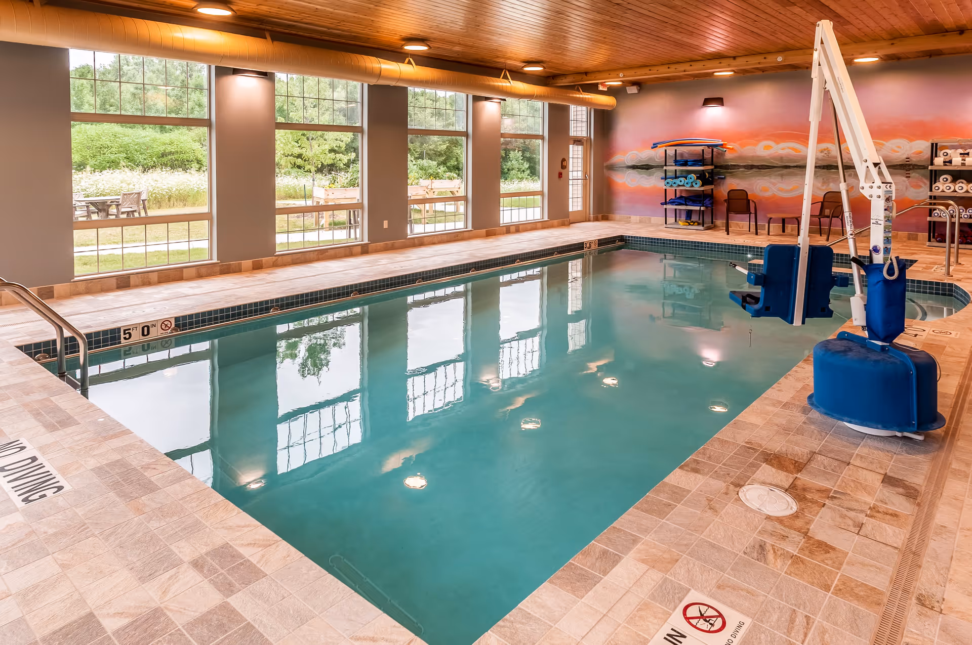 Indoor swimming pool with tiled floor and walls, large windows showing greenery outside, pool lift for accessibility, and shelves with rolled towels and pool equipment.