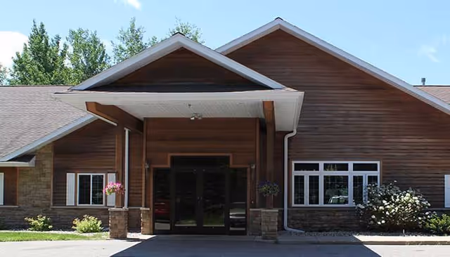 Front exterior view of a single-story assisted living facility building with a covered entrance, wooden siding, stone accents, and windows. There are some bushes and trees around the building under a clear blue sky.