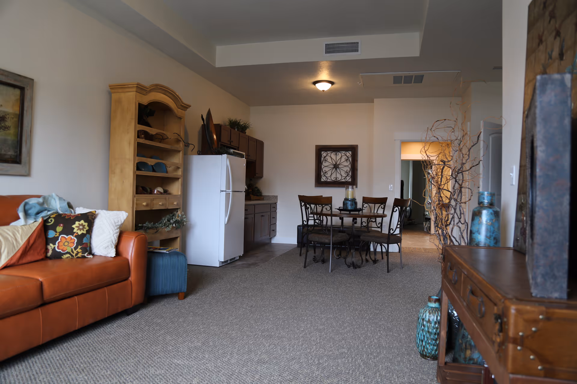Interior view of a senior living facility room featuring a brown leather couch with colorful pillows on the left, a wooden shelving unit with shoes, a white refrigerator, a small dining table with four chairs, and decorative vases and branches on the right side. The room has carpeted flooring and neutral-colored walls.
