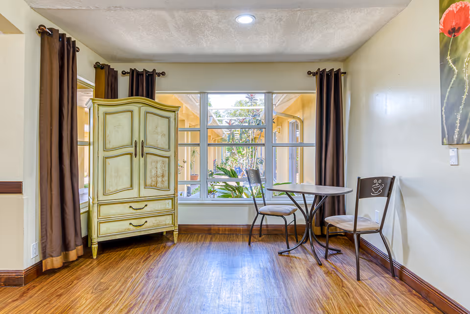 Sunlit interior nook with a round bistro table and two chairs by a large window and a vintage armoire on wood floors.
