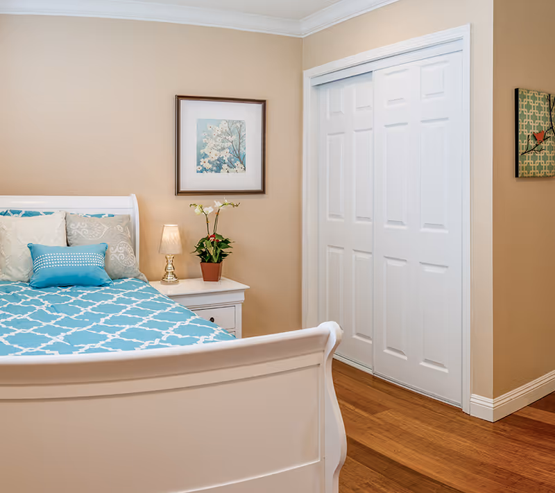 A cozy bedroom with a white wooden bed frame and a blue and white patterned bedspread. The bed has several pillows in shades of beige and blue. Next to the bed is a white nightstand with a small lamp and a potted plant. The walls are beige with two framed artworks hanging, one above the nightstand and one on the adjacent wall. There is a white sliding closet door and wooden flooring.