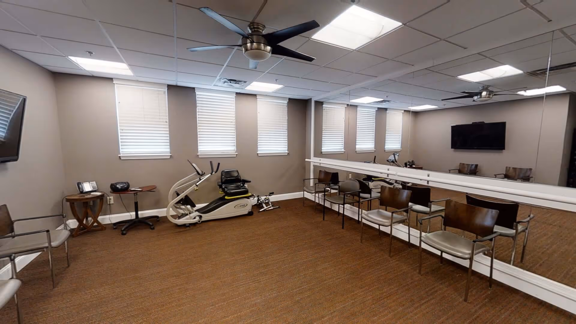 A fitness room with exercise equipment including a recumbent stepper bike, several chairs arranged along the walls, a large wall mirror, a ceiling fan, and a wall-mounted television. The room has beige walls, carpeted floor, and several windows with blinds.
