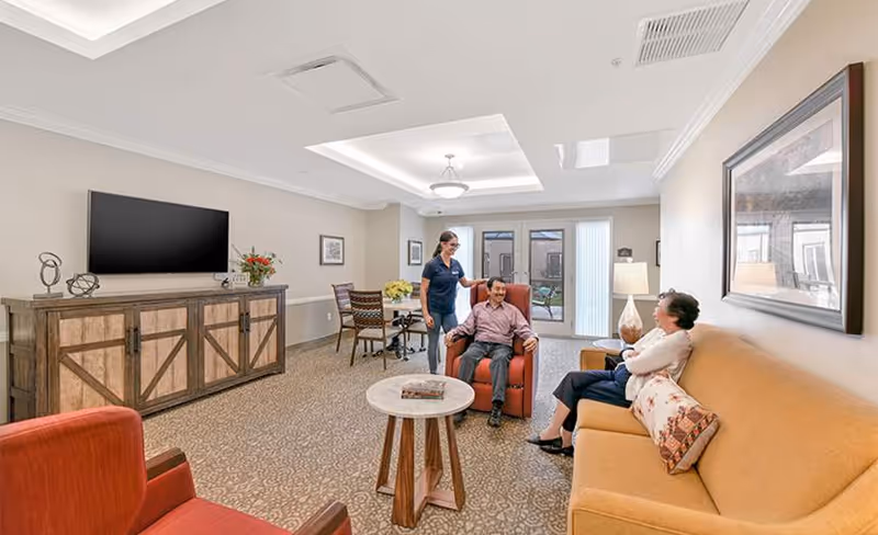 A bright memory care living room with sofas, armchairs, a TV, and a caregiver assisting two seated residents.