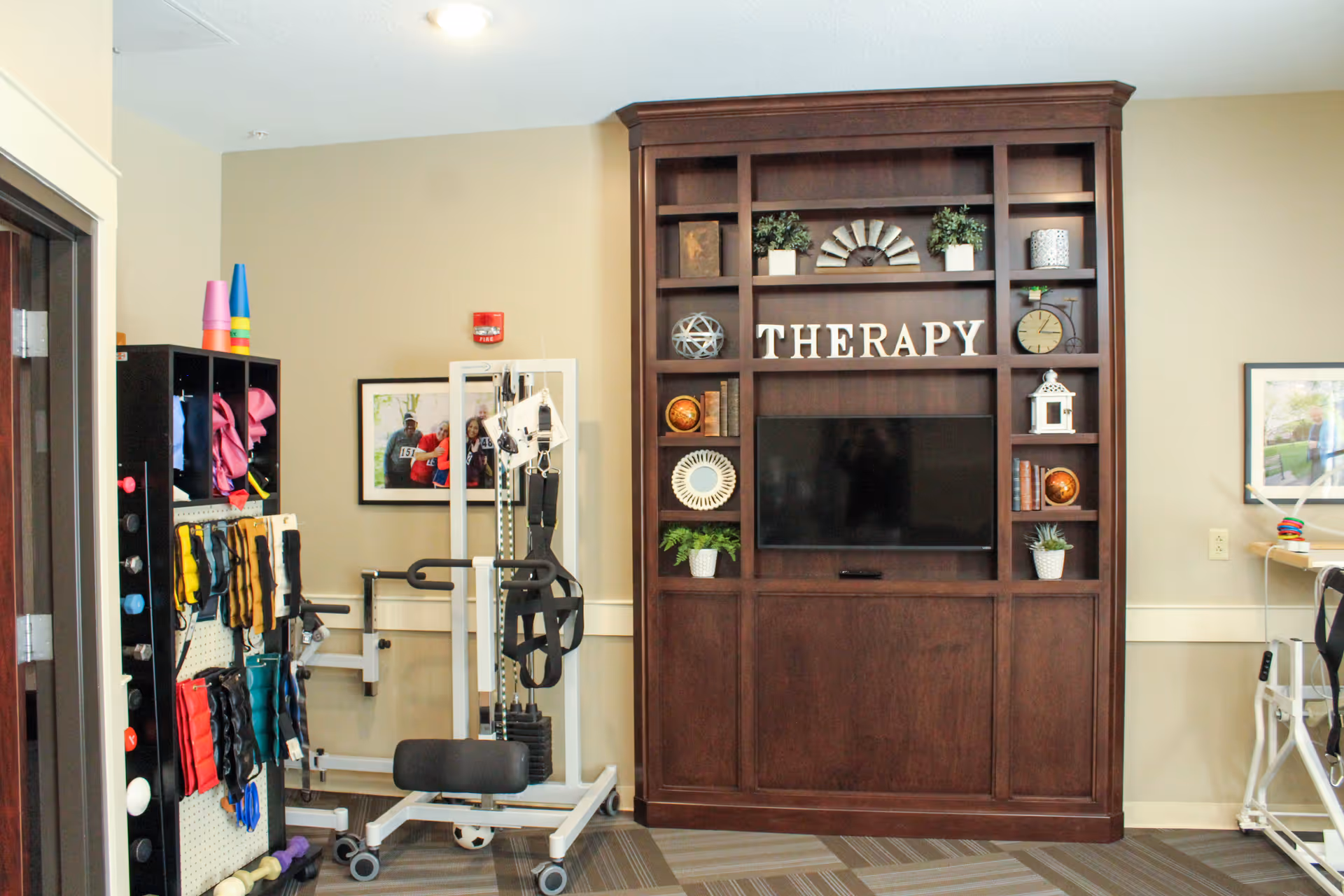 A therapy room with exercise equipment including resistance bands, weights, and a pulley system. There is a large wooden shelving unit with decorative items and the word 'THERAPY' displayed above a flat-screen TV. The room has beige walls and framed pictures on the walls.