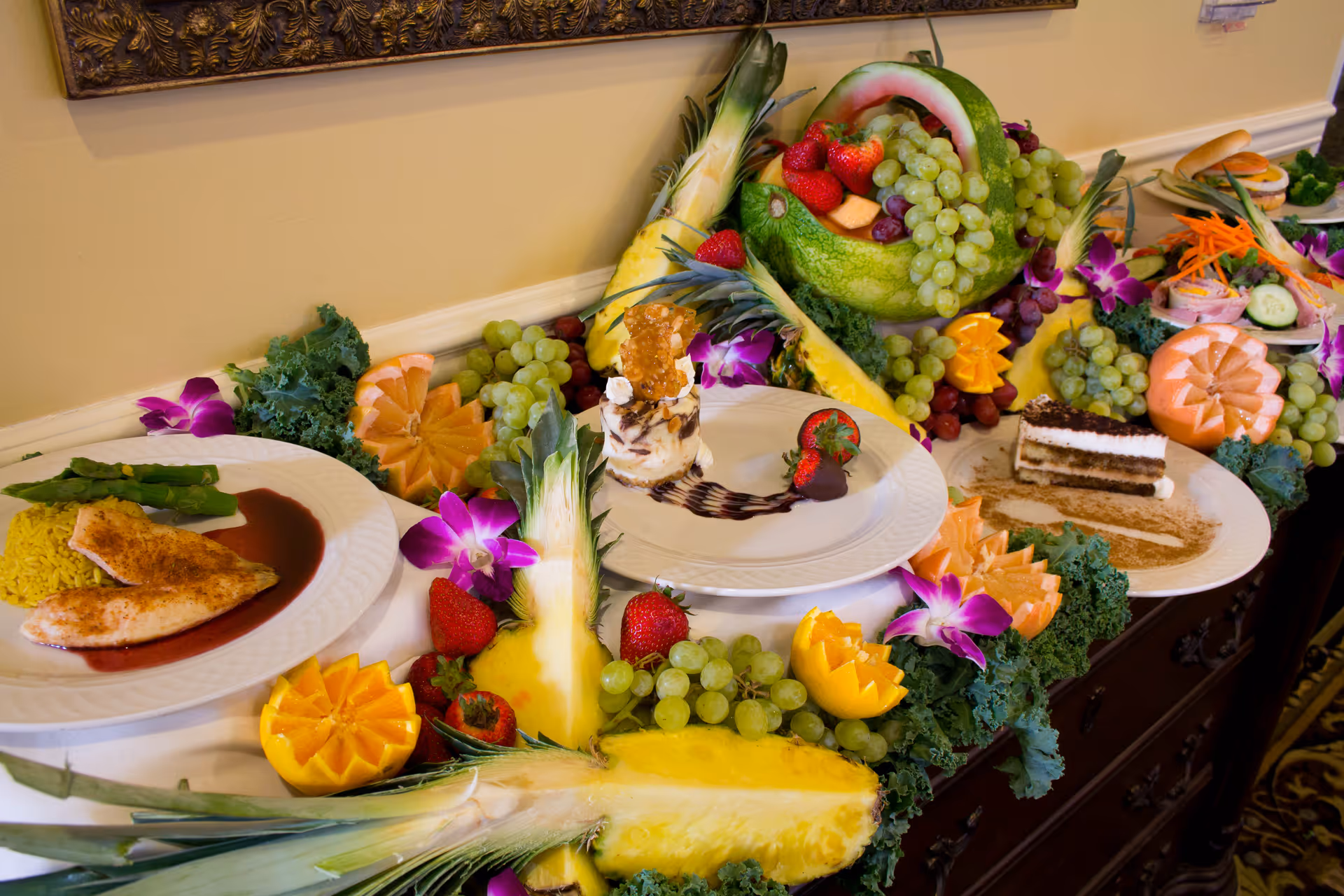 A buffet table with an assortment of plated foods including grilled fish with asparagus and rice, a dessert with strawberries and chocolate drizzle, and a slice of layered cake. The table is decorated with an elaborate fruit arrangement featuring pineapple, grapes, strawberries, carved melons, and purple flowers.