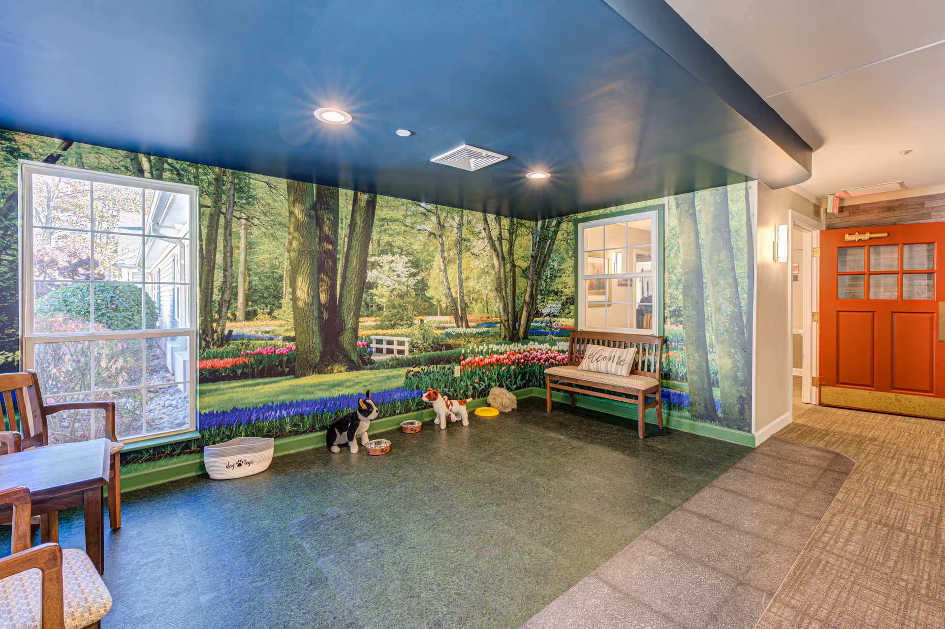 Indoor room with a large mural of a forest and garden scene covering two walls. The room has a green floor, a wooden bench with a 'Welcome' pillow, a small table with chairs, and several dog bowls and dog toys on the floor. There is a red door in the background and a window on the mural wall.