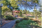 Paved pathway winding through a landscaped garden with trees, shrubs, and a covered gazebo on a sunny day.