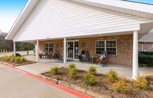 Covered front porch area of a brick building with white columns and a white roof overhang. There are several chairs and potted plants arranged along the porch. A small garden bed with shrubs is in front of the porch, and a driveway with a red curb runs alongside it.