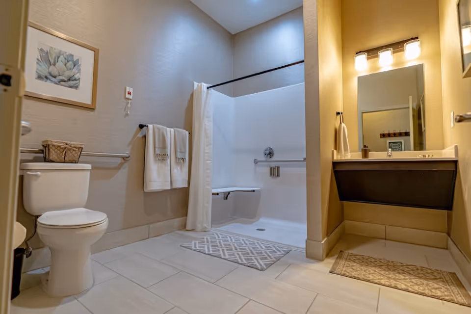 A clean and modern bathroom featuring a white toilet with a basket on top, a towel rack with two white towels, a walk-in shower with a fold-down seat and grab bars, a white shower curtain, a beige tiled floor with two patterned rugs, and a vanity with a sink, mirror, and three light fixtures above.