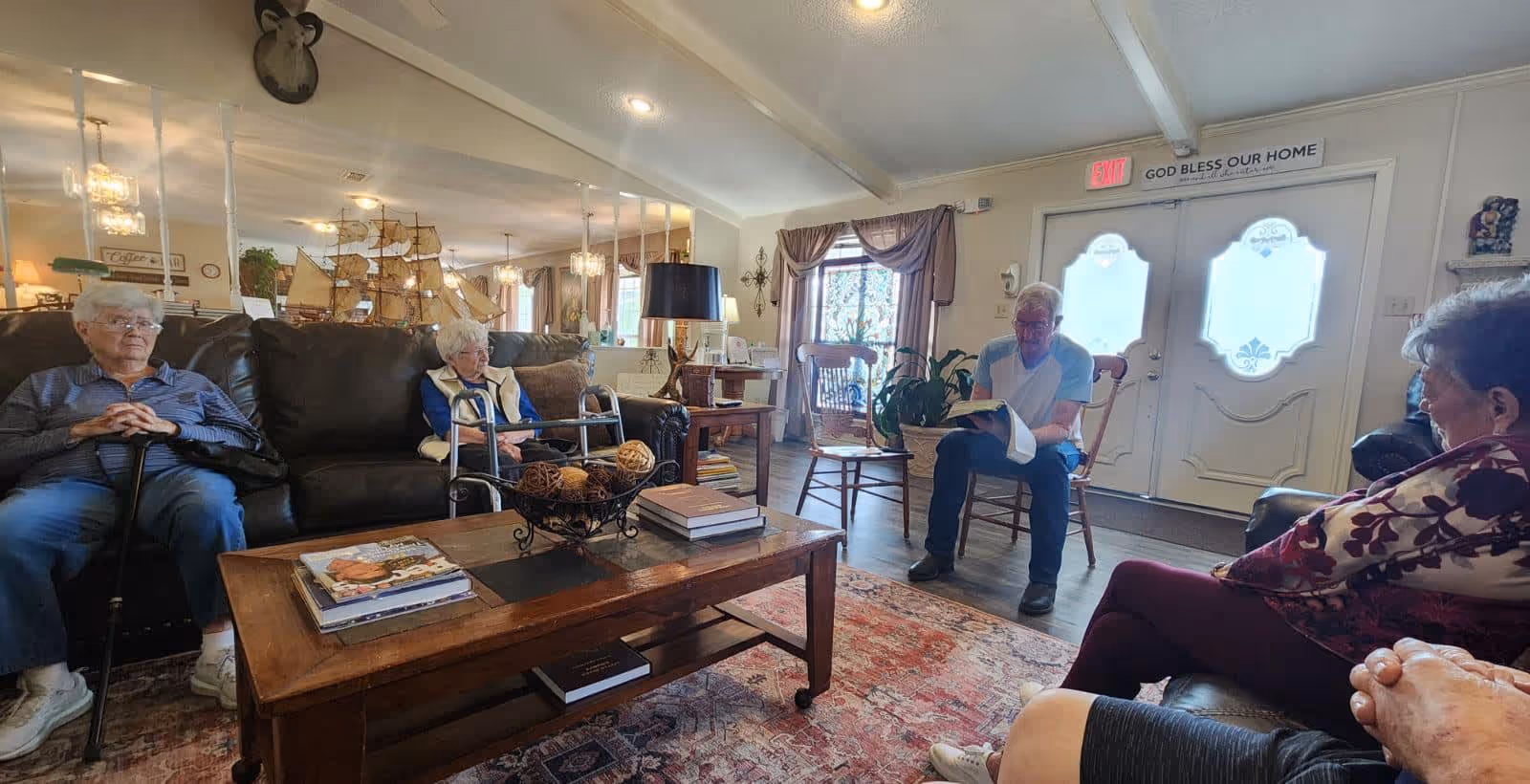 A cozy living room in Pinecrest Retirement Lodge with several elderly residents seated on sofas and chairs arranged around a wooden coffee table. The room features warm lighting, decorative items including a large model ship, a patterned rug, and a sign above the door that reads 'GOD BLESS OUR HOME'.