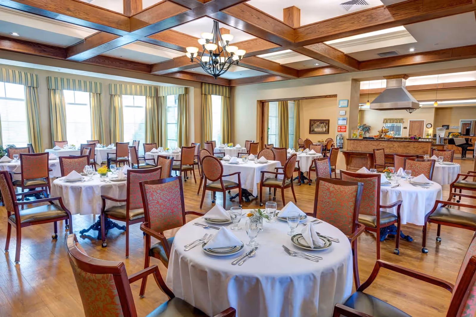 Spacious dining room with round tables set with white tablecloths and place settings, wooden chairs, and a coffered ceiling with chandelier.