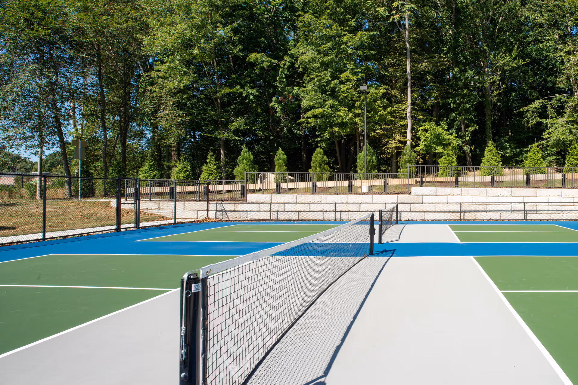 Outdoor tennis court with a net in the center, surrounded by a black chain-link fence and trees in the background under a clear blue sky.