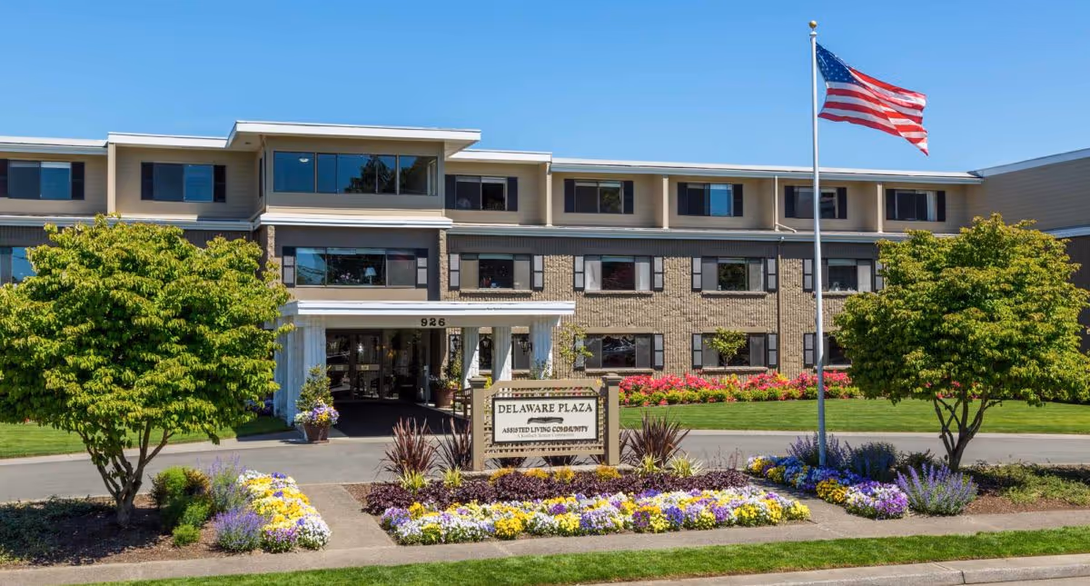 Front exterior view of Delaware Plaza Assisted Living Community building with a well-maintained garden featuring colorful flowers, two trees, and an American flag on a flagpole in front of the entrance.