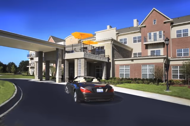 Exterior view of a senior living facility named All Seasons West Bloomfield with a covered entrance. A black convertible car is parked on the driveway in front of the building. The building has multiple windows, balconies with yellow umbrellas, and well-maintained landscaping under a clear blue sky.