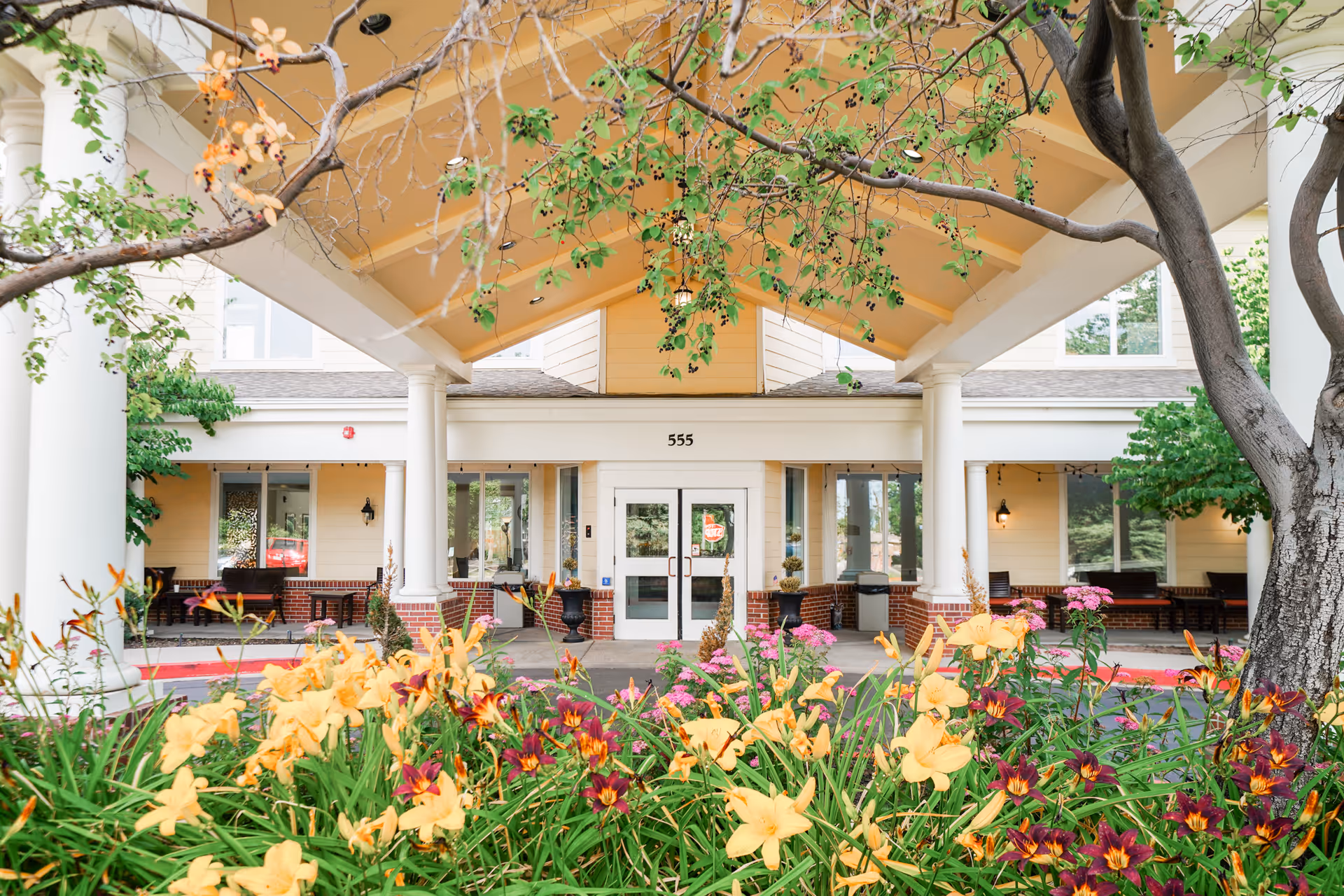Entrance of a senior living facility with a covered drop-off area supported by white columns. The building is light yellow with white trim and has large windows. In the foreground, there are colorful flowers including yellow and purple blooms, and a tree with green and some orange leaves. The number 555 is displayed above the double glass doors.