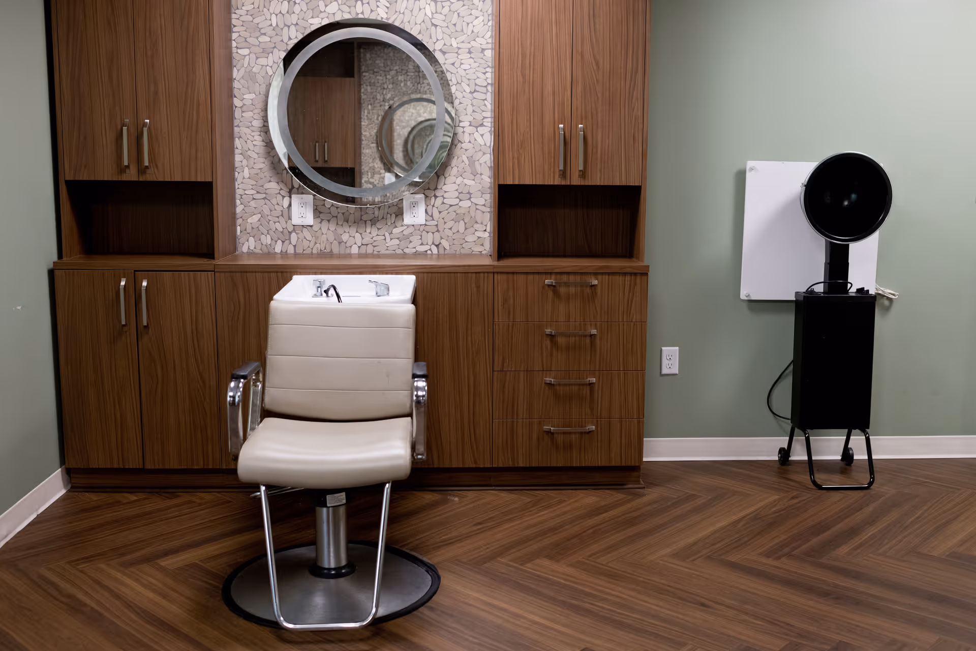 A salon area with a beige salon chair in front of a wooden cabinet with drawers and shelves. Above the cabinet is a round mirror mounted on a pebble-textured wall section. To the right of the chair is a black hair dryer on a stand. The floor has a wood-patterned design and the walls are painted light green.