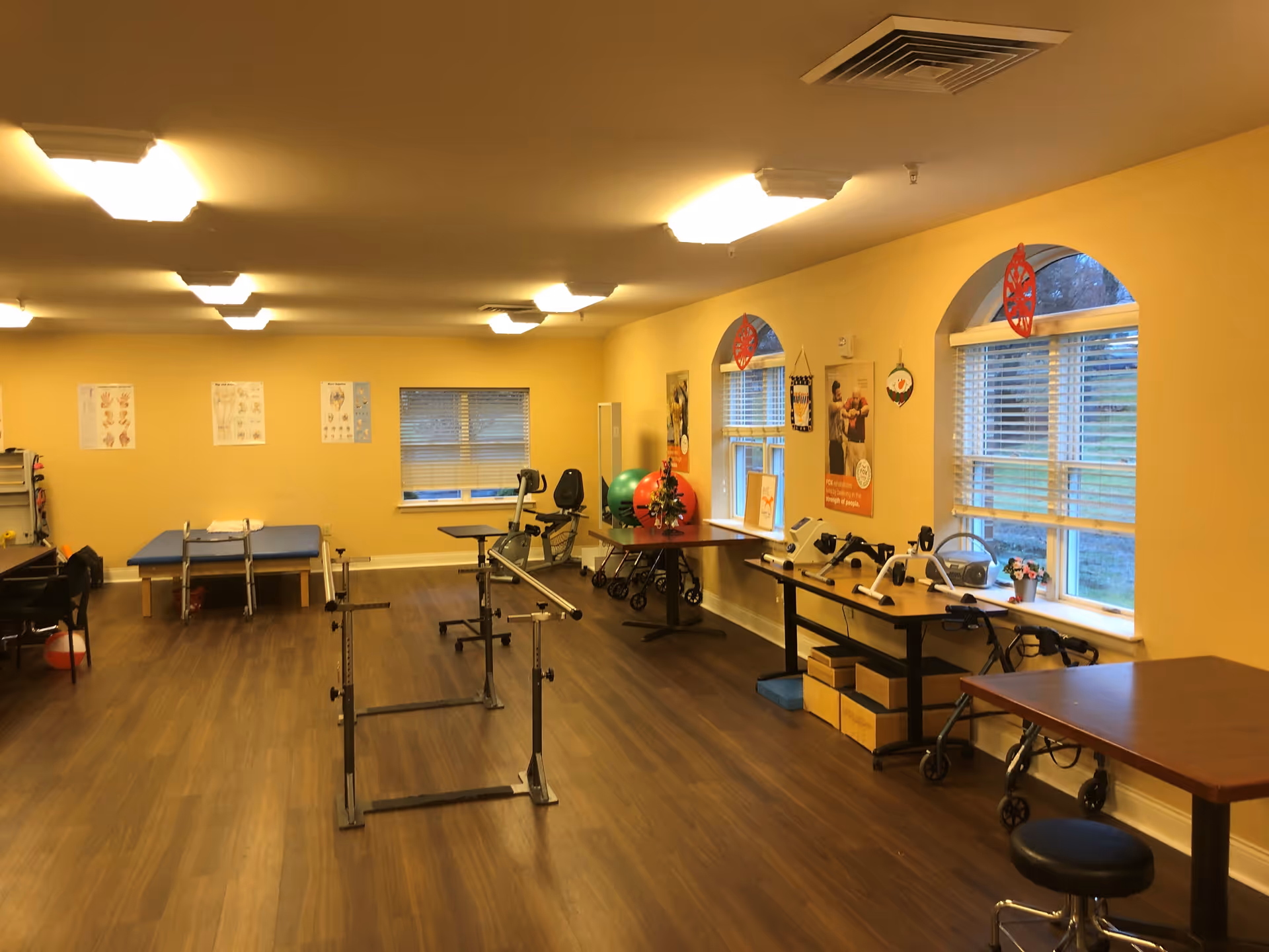 A spacious rehabilitation/exercise room with parallel bars, therapy tables, exercise equipment and large windows.
