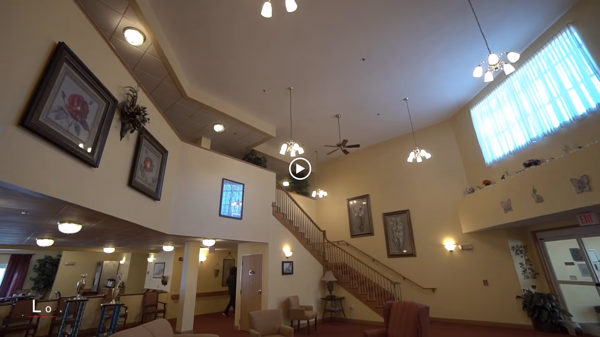Interior view of a senior living facility lobby with a staircase leading to an upper level. The walls are painted light yellow and decorated with framed floral artwork. There are several ceiling lights and a large window with sheer curtains allowing natural light to enter. Seating areas with chairs and a small table are visible, along with plants and decorative items on ledges.