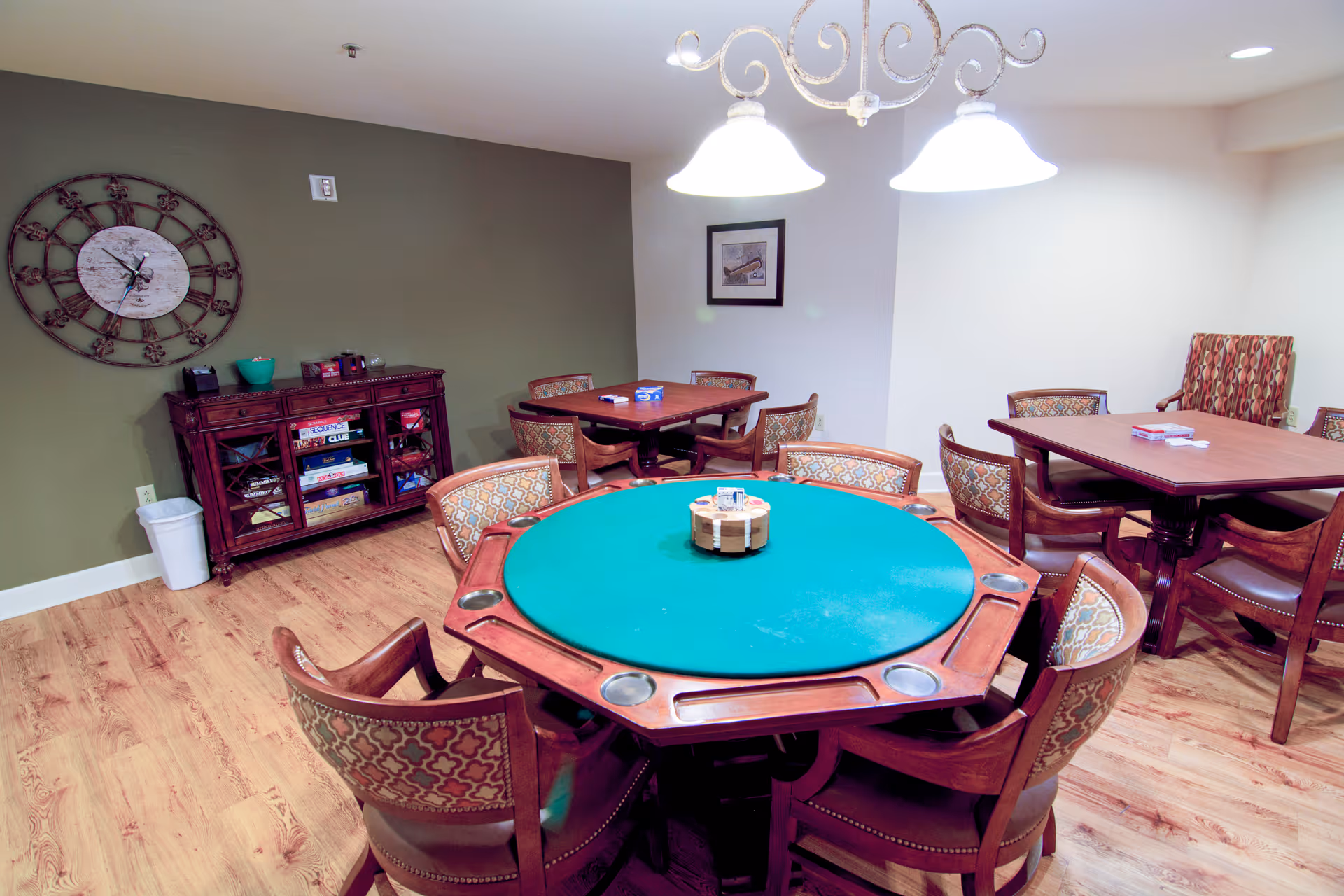 Interior game room with a green-felt poker table surrounded by wooden upholstered chairs, additional card tables, a sideboard and overhead lights.