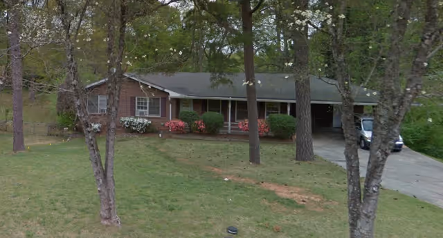 A single-story brick house with a covered front porch and a driveway on the right side. The yard has green grass, several trees with white blossoms, and bushes with pink and white flowers near the house.