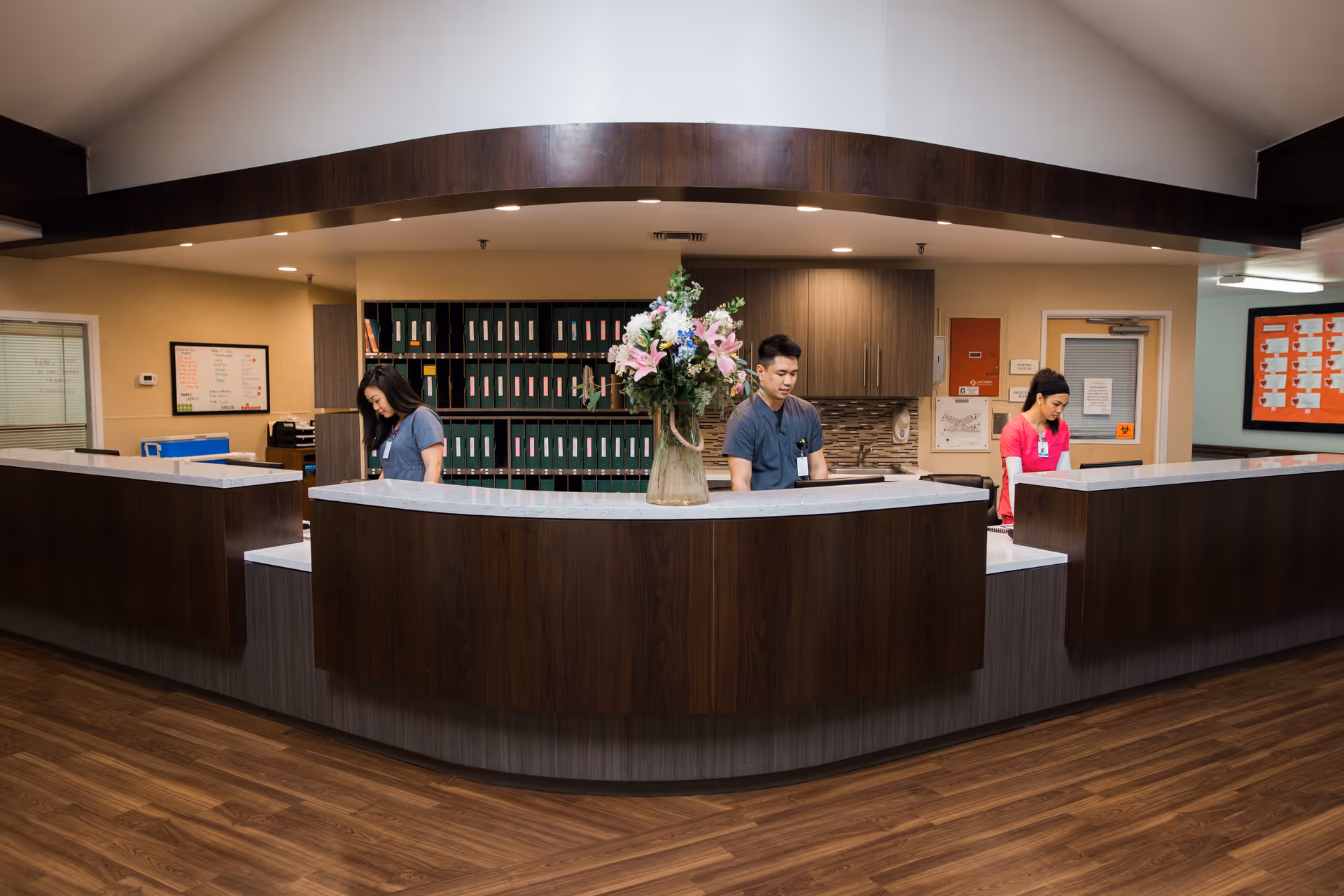 Reception area in a healthcare facility with a curved wooden desk and three staff members working behind it. There is a large vase with flowers on the desk, shelves with organized files in the background, and a bulletin board on the wall.