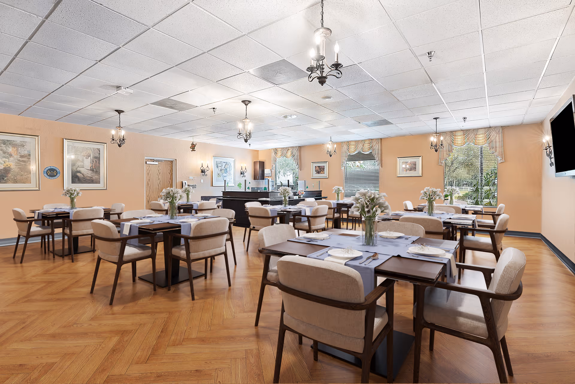 Bright dining room with multiple tables set with plates and floral centerpieces, upholstered chairs, chandeliers, and large windows.