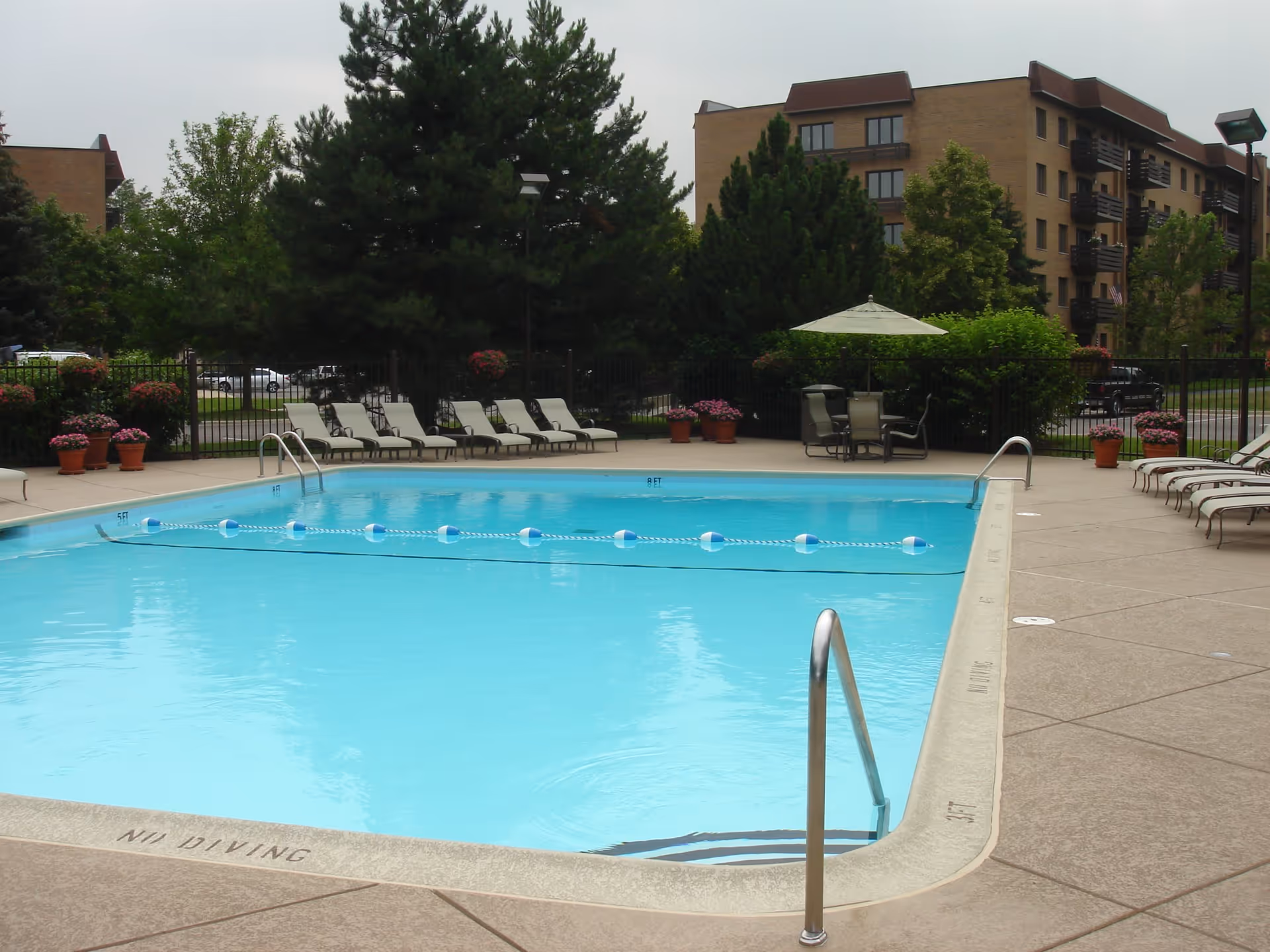 Outdoor swimming pool with lounge chairs, potted plants, and apartment buildings in the background.