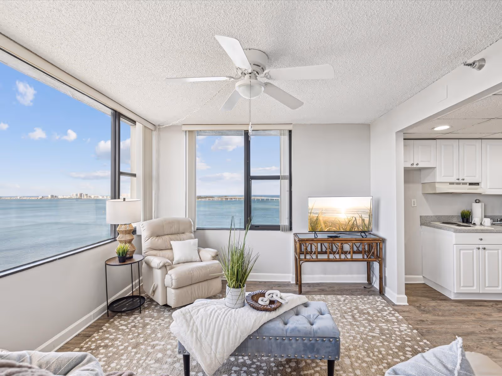 Bright living room with large windows offering a panoramic view of a body of water and distant buildings. The room features a white ceiling fan, a beige recliner chair next to a small round side table with a lamp and plant, a blue tufted ottoman with a white throw blanket and decorative items, a TV on a wooden stand, and an open view into a white kitchen area.