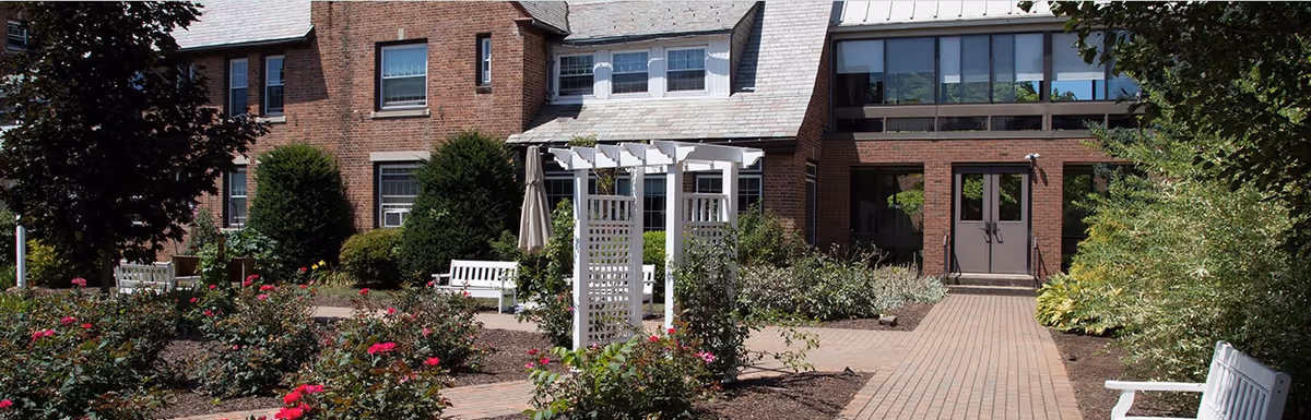 Brick senior living building with a paved pathway through a rose garden, white benches, and a pergola leading to the entrance.