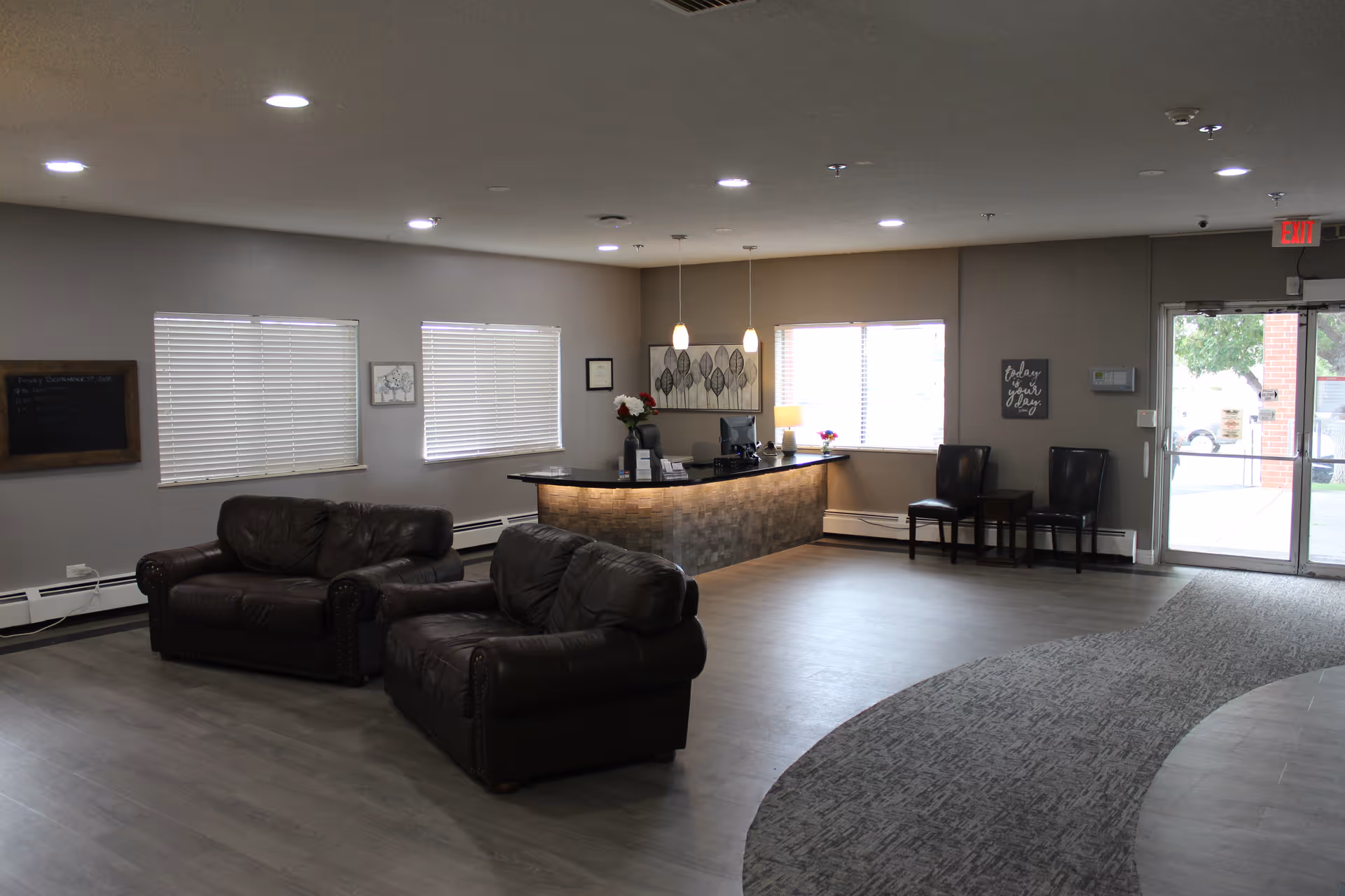A spacious reception area with two dark brown leather sofas, a curved reception desk with a computer and decorative items, three chairs against the wall near a glass door exit, and windows with blinds letting in natural light. The floor is a combination of wood and carpet, and the walls are painted light gray.