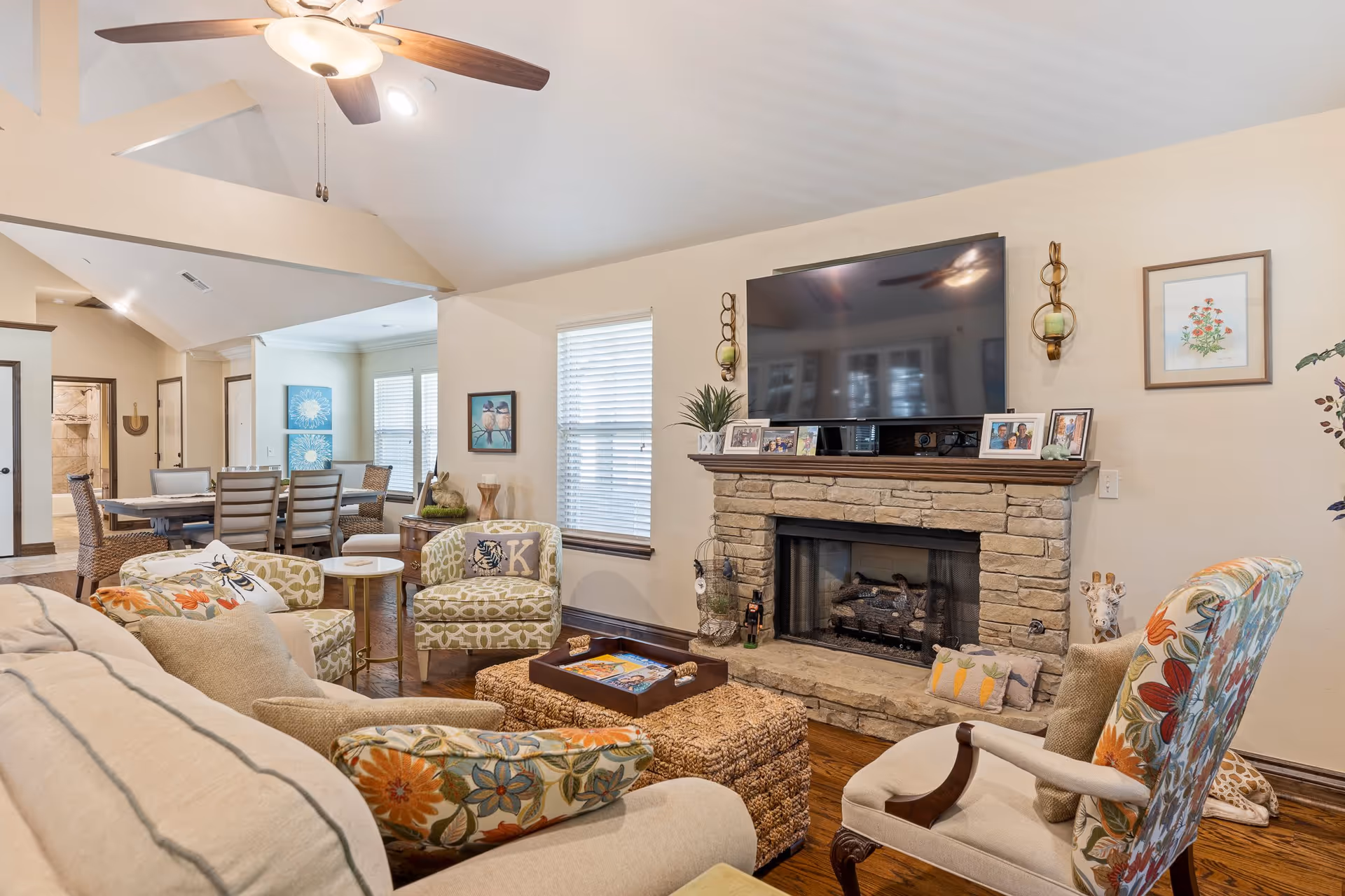 A cozy living room with a beige sofa adorned with floral and striped cushions, two patterned armchairs, and a woven ottoman in the center. A large flat-screen TV is mounted above a stone fireplace with a wooden mantle displaying family photos and decorative items. The room has hardwood floors, a ceiling fan with lights, and windows with blinds. In the background, a dining area with a table and chairs is visible.
