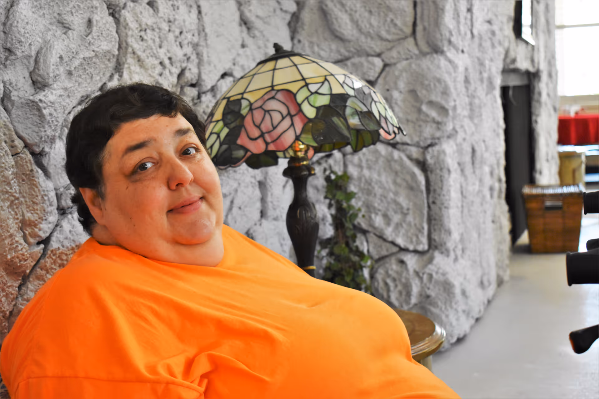 A person wearing an orange shirt is sitting indoors against a textured stone wall. Behind them is a decorative lamp with a stained glass shade featuring a floral design. The setting appears to be a cozy interior space with some furniture and decor visible in the background.