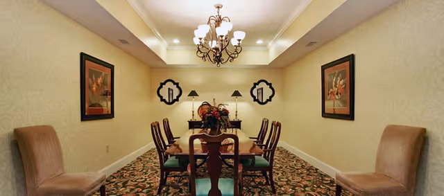 A formal dining room with a long wooden table surrounded by eight chairs. The room features patterned carpet, beige wallpaper with a subtle design, two framed floral paintings on the side walls, two decorative mirrors on the far wall, a chandelier hanging from the ceiling, and a floral centerpiece on the table.
