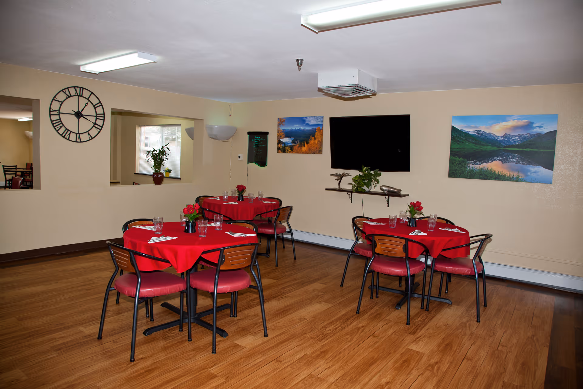 Dining room with round tables covered in red tablecloths, chairs, a wall-mounted TV, and landscape artwork.