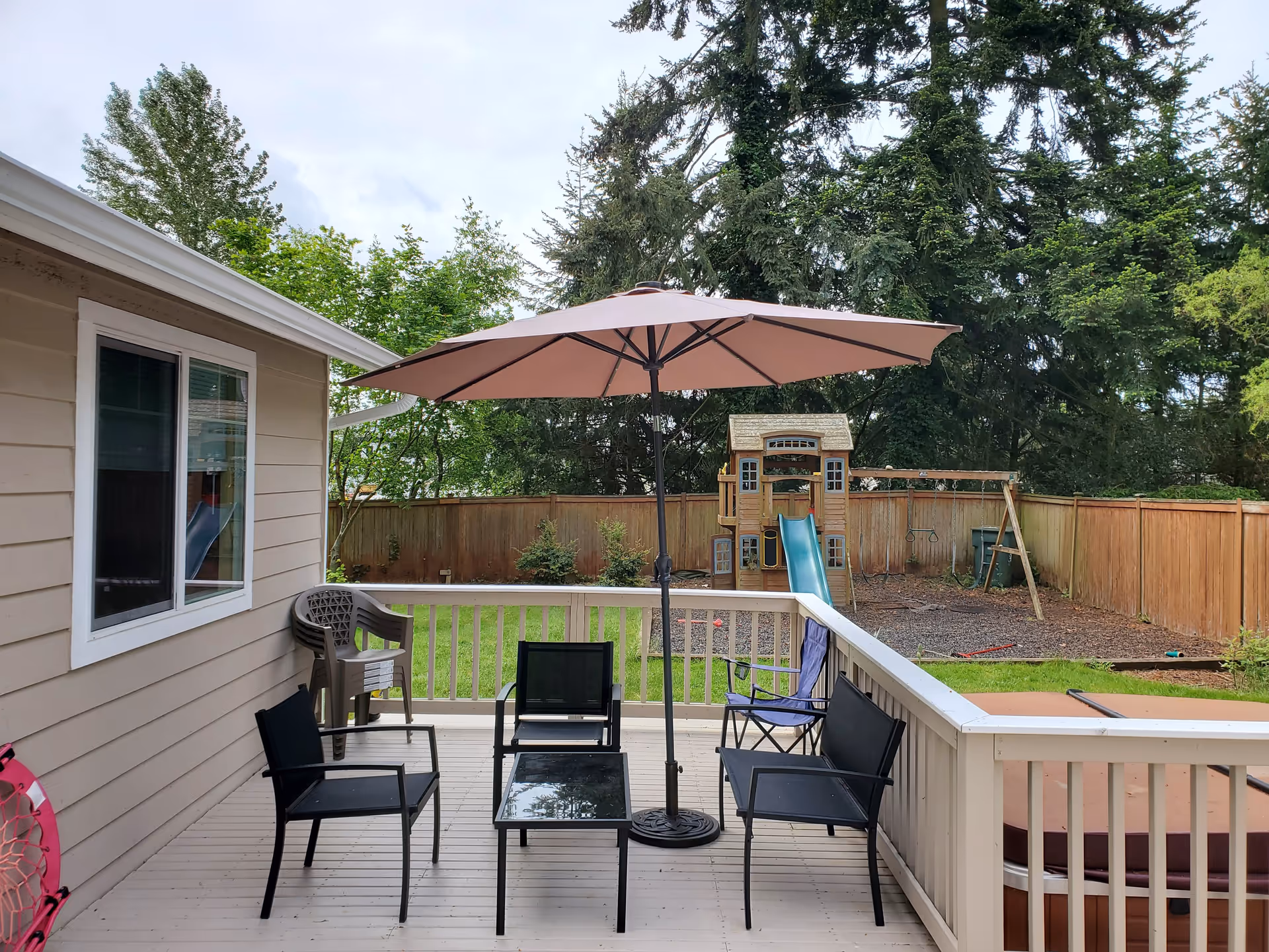 Outdoor patio area with black chairs and a glass-top table under a large beige umbrella. The patio is attached to a beige house with a window. In the background, there is a fenced backyard with green grass, trees, and a wooden playset with a slide and swings.