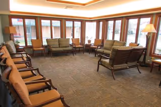 A spacious living room with large windows along two walls allowing natural light to fill the space. The room is furnished with multiple cushioned chairs and sofas arranged around the perimeter, with a carpeted floor and warm lighting from ceiling fixtures and a floor lamp.