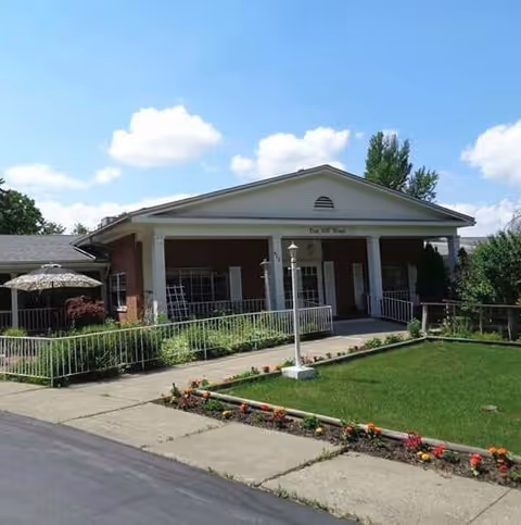 Front exterior view of Oak Hill Manor, a single-story brick building with white columns supporting a triangular pediment. There is a well-maintained lawn with flower beds and a paved walkway leading to the entrance. The sky is blue with some clouds.