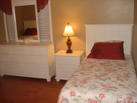 A bedroom featuring a floral bedspread, nightstand with lamp, and a white dresser with mirror.