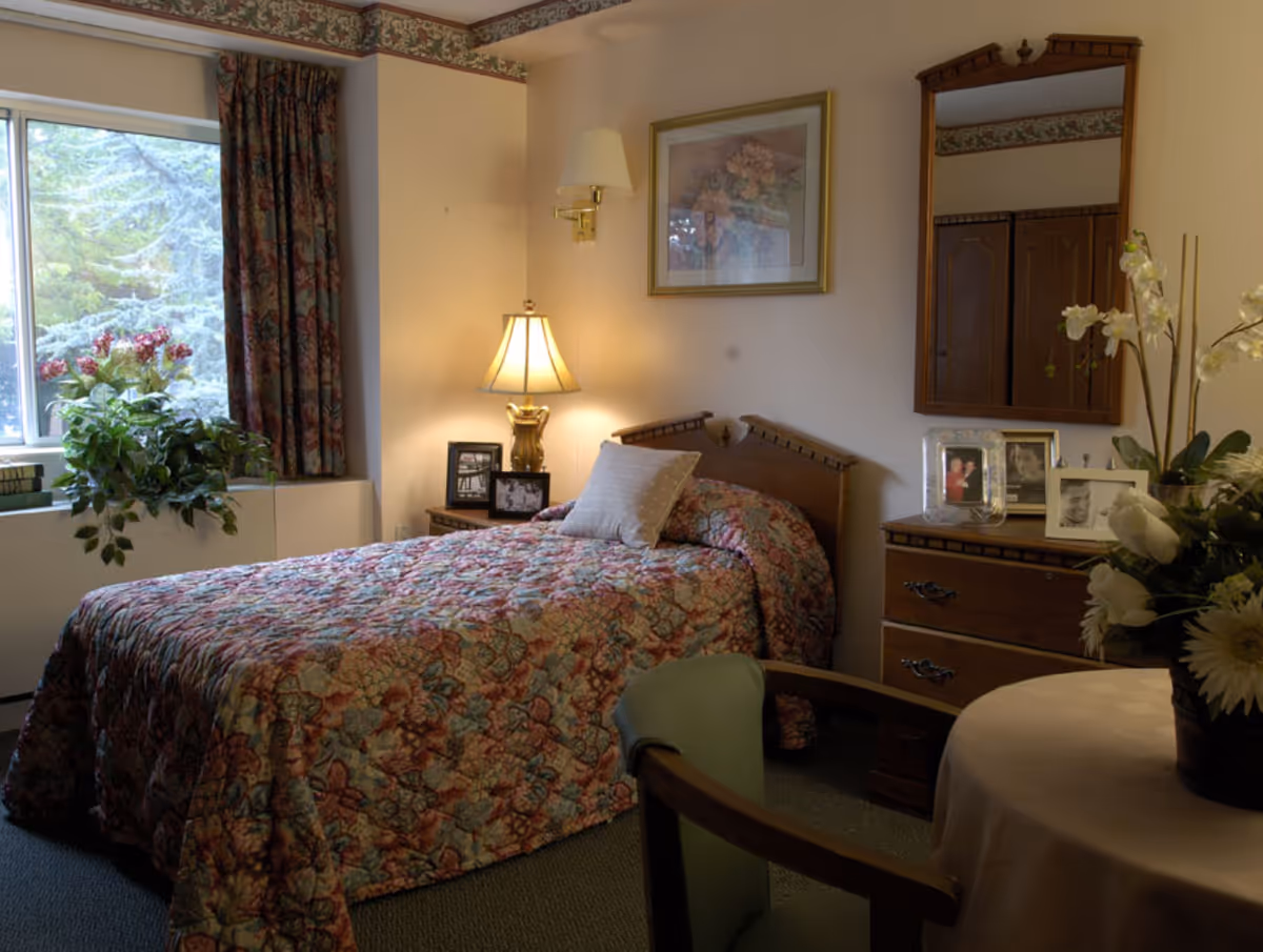 A cozy senior bedroom featuring a single floral bed, bedside lamp and table, dresser with framed photos, and a window with plants.