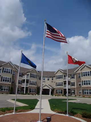 Front exterior of Parkside Village senior living building with three flagpoles—an American flag centered—and a walkway leading to the main entrance.